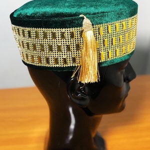 Puede incluir: Un sombrero de terciopelo verde con ribete dorado y una borla dorada. El sombrero se exhibe en una cabeza de maniquí negra. El sombrero tiene un ribete dorado rectangular alrededor de la base y una borla dorada colgando del lateral.