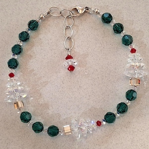 May include: A festive bracelet featuring dark green faceted beads, clear crystal tree charms, and small red accents. The bracelet has a silver-tone chain and clasp. A small charm with red and clear beads hangs from the chain.