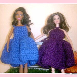 May include: Two dolls wearing handmade crochet dresses. One doll is wearing a blue dress, and the other is wearing a purple dress. The dresses have a halter-style top and a full skirt. The dolls are standing next to each other. The image is framed with a pink border.