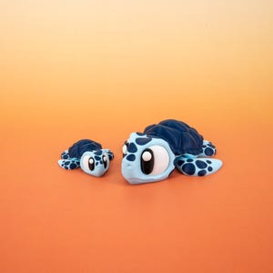 May include: Two blue and black sea turtle figurines with large eyes. The larger turtle is facing the left and the smaller turtle is facing the right. Both turtles are on a light orange background.