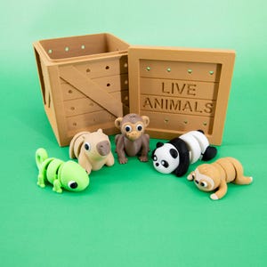 Puede incluir: Una caja de madera con las palabras "LIVE ANIMALS" impresas en el lateral, rodeada de gomas de borrar con forma de animales. Las gomas incluyen un lagarto verde, un carpincho beige, un mono marrón, un panda y un perezoso beige.