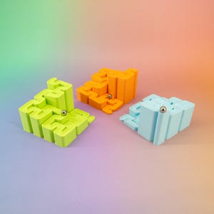 Könnte beinhalten: Drei farbenfrohe Labyrinth-Puzzles in Limettengrün, Orange und Hellblau. Jedes Puzzle hat eine einzigartige Form und eine kleine silberne Kugel im Inneren. Der Hintergrund hat einen Farbverlauf, der eine lebendige Szene erzeugt.