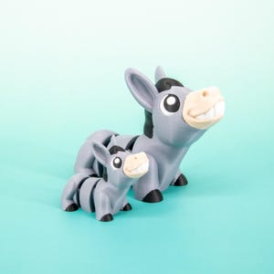May include: Two gray donkey figurines, one large and one small, are displayed against a light blue background. The donkeys have black manes, ears, and hooves, with smiling faces and tan noses. The larger donkey stands upright, while the smaller one is in a seated position.