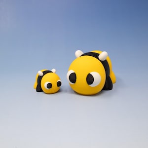 May include: Two yellow and black bumblebees with white eyes. The larger bee is facing the camera, while the smaller bee is facing to the right. Both bees are made of clay.