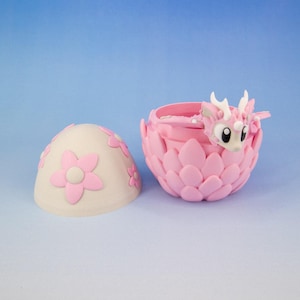 May include: A pink and white egg-shaped container with a pink flower design. The container opens to reveal a pink dragon figurine inside.