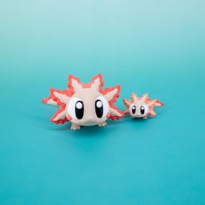 May include: Two axolotl figurines, one larger than the other, are made of a light pink and orange material. The larger axolotl has large black eyes and a wide smile. The smaller axolotl has smaller black eyes and a smaller smile.