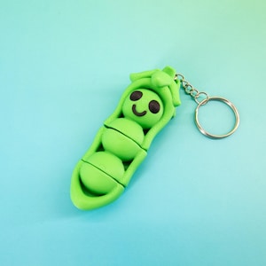 May include: A green pea pod keychain with three peas inside. The peas have black eyes and a smiling face. The keychain is made of a smooth material and has a silver-colored keyring attached.