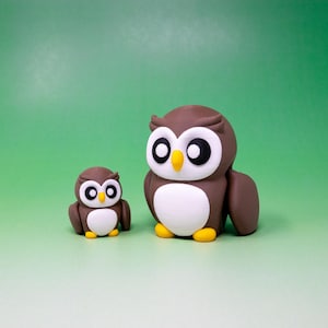 Pode incluir: Duas estatuetas de coruja marrons com barrigas brancas e bicos amarelos. A coruja maior está em pé com as asas abertas, enquanto a coruja menor está sentada com as asas fechadas.