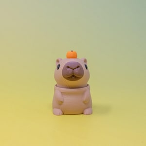 Puede incluir: Una figurita de capibara marrón y beige con una pequeña calabaza naranja en la cabeza. La figurita está sentada sobre una superficie amarilla.