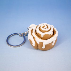 May include: A miniature cinnamon roll keychain with white icing detail. The keychain is made of a light brown material and has a silver keyring attached. The background is a solid light blue.