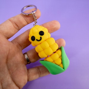 Puede incluir: Un llavero con forma de mazorca de maíz amarilla con una cara sonriente, ojos negros y una cáscara verde. El llavero está unido a un llavero plateado. El maíz está hecho de un material blando.