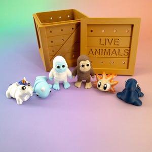May include: A wooden crate with the words "LIVE ANIMALS" printed on the side, alongside six colourful animal figurines. The figurines include a unicorn, a dolphin, a yeti, a monkey, a sun-like creature, and a seal.