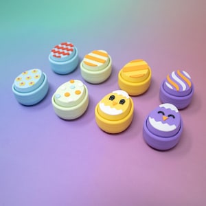 Puede incluir: Ocho recipientes coloridos en forma de huevo de Pascua con diferentes diseños, que incluyen lunares, rayas y una cara de pollito. Los recipientes están hechos de plástico y tienen una combinación de colores azul claro, amarillo, morado y verde.