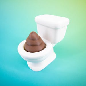 Pode incluir: Uma sanita branca com um emoji de cocó castanho esculpido no interior. A sanita está colocada sobre um fundo gradiente de verde-azulado e verde. A sanita é um artigo de novidade.