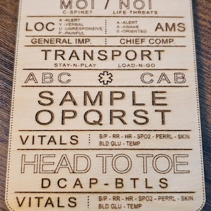 May include: Wooden engraved medical assessment card with text including "Scene Safety", "MOI/NOI", "ABC", "Head to Toe", and "Vitals". The card is rectangular with rounded corners and is made of light-colored wood.