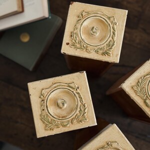 May include: A set of square wooden canisters with cream-coloured lids featuring a raised circular medallion and leaf-like detailing. The lids have a distressed finish with visible brown wood tones showing through the paint on the edges.