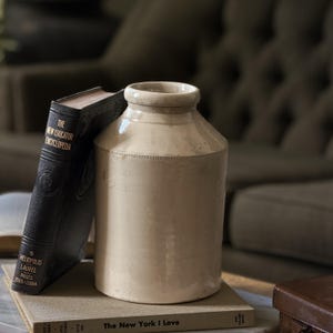 May include: A cylindrical cream-coloured ceramic crock with a thick rim and a smooth glazed finish, positioned next to a black hardcover book titled The New Educator Encyclopedia and resting on a beige book titled The New York I Love.