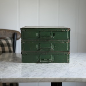 May include: A stack of three rectangular forest green metal storage drawers. Each drawer features a flat front panel with a central horizontal handle and a small integrated label holder frame. The metal surfaces show signs of wear and patina.