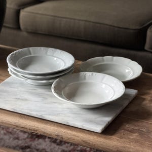 May include: A set of four white ceramic bowls with a wide, scalloped rim featuring a subtle embossed pattern. The bowls have a smooth, glossy finish and a shallow, rounded basin. Three bowls are stacked together, while one sits separately.