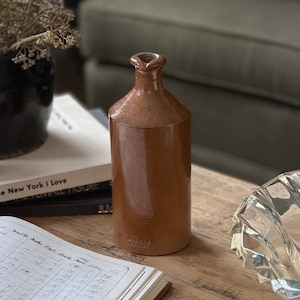 Peut inclure: Une bouteille cylindrique en céramique brune dotée d'un col étroit et d'un petit bec verseur. La surface présente une finition lisse et émaillée avec de légères taches. Un petit tampon en relief est visible près de la base de la bouteille.