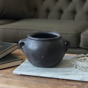 May include: A dark, round, ceramic pot with two small handles. The pot is a matte black colour and sits on a white marble tray. The background includes a grey sofa and a wooden table.