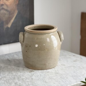 May include: A beige ceramic pot with two side handles and horizontal etched lines. The pot has a wide opening and a slightly lighter rim. The pot is sitting on a marble surface.
