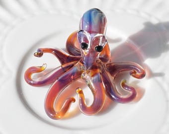 May include: A colorful glass octopus figurine with a blue head, purple and orange body, and black eyes. The tentacles curl gracefully. The figurine is displayed on a white plate.