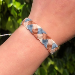 May include: Woven friendship bracelet in a repeating pattern of blue, tan, and brown threads. The bracelet is on a wrist, with a braided tie closure. The background is blurred green foliage.