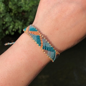 Può includere: Un braccialetto in macramè intrecciato nei colori verde acqua, arancione e beige, con dettagli dorati. Il braccialetto è indossato su un polso, mostrando l'intricato motivo e la combinazione di colori. Lo sfondo è una vista sfocata di fogliame verde e acqua.