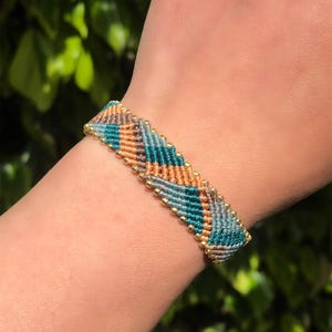 May include: A macrame bracelet with a chevron pattern in teal, orange, and grey. The bracelet is accented with small gold beads along the edges. The bracelet is approximately 2 cm wide.