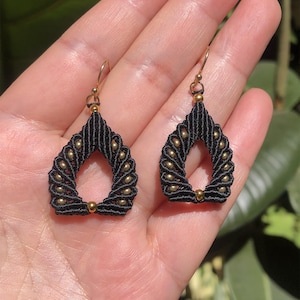 Peut inclure: Une paire de boucles d'oreilles en forme de goutte composées de cordon macramé noir tissé en une structure rigide. Chaque boucle est ornée de petites perles métalliques dorées rondes insérées sur les bords, suspendues à des crochets dorés.