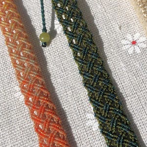 K&ouml;nnte beinhalten: Zwei geflochtene Armb&auml;nder mit diagonalem Gittermuster. Ein Armband ist in leuchtendem Orange mit subtilem metallischem Glanz gehalten, w&auml;hrend das andere in tiefem Waldgr&uuml;n mit dunklen F&auml;den und einer kleinen, runden, hellgr&uuml;nen Perle versehen ist.