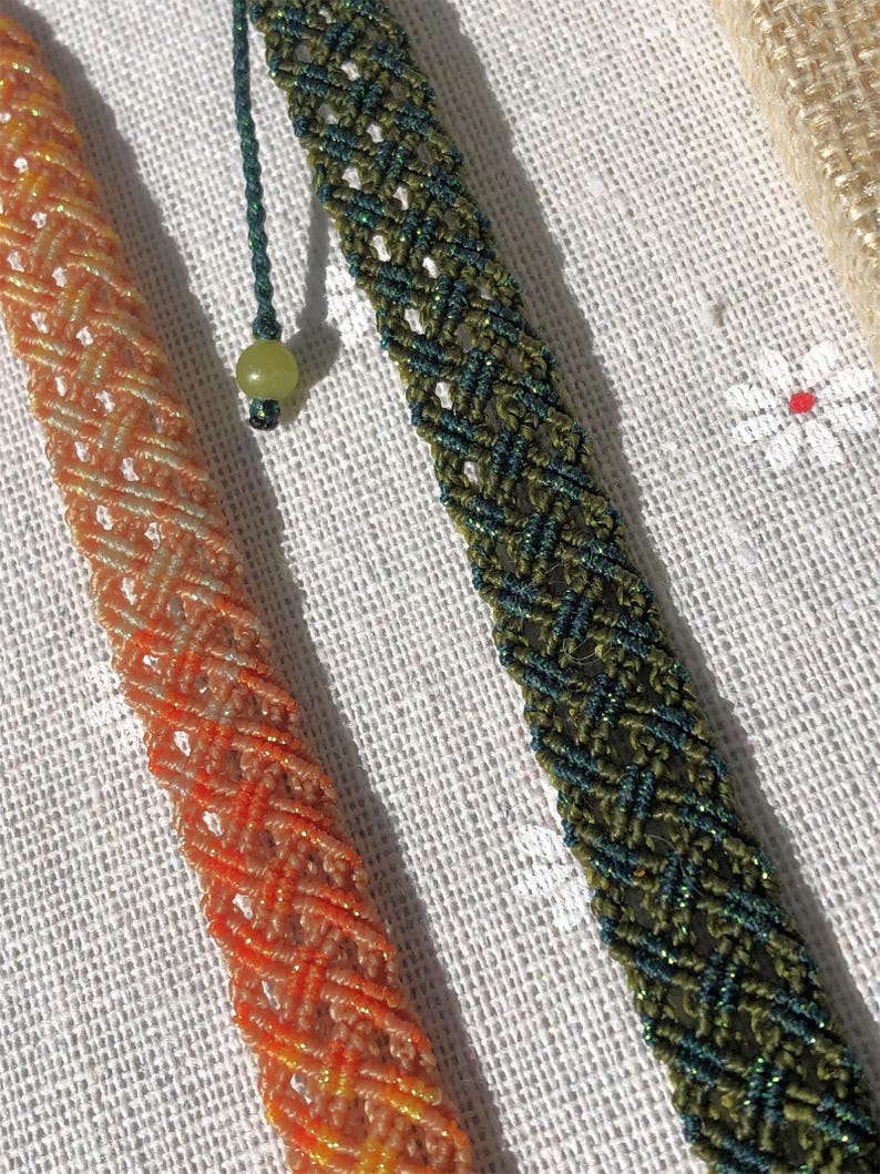 K&ouml;nnte beinhalten: Zwei geflochtene Armb&auml;nder mit diagonalem Gittermuster. Ein Armband ist in leuchtendem Orange mit subtilem metallischem Glanz gehalten, w&auml;hrend das andere in tiefem Waldgr&uuml;n mit dunklen F&auml;den und einer kleinen, runden, hellgr&uuml;nen Perle versehen ist.
