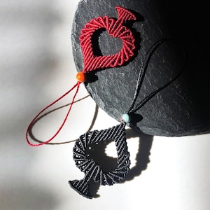 May include: Two macrame pendants, one red heart-shaped and one black spade-shaped, each with a small bead and a cord. The red pendant has an orange bead, and the black pendant has a turquoise bead. The pendants are displayed on a dark gray stone.
