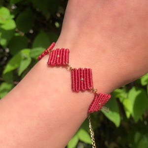 Könnte beinhalten: Ein rotes Makramee-Armband mit einer Goldkette und Akzenten. Das Armband hat drei rechteckige gewebte Abschnitte, die durch kleine Goldketten verbunden sind. Das Armband wird an einem Handgelenk getragen, mit einem grünen, blättrigen Hintergrund.