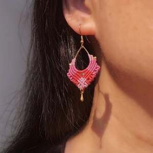 May include: A handmade macrame earring with a gold-colored hook and a teardrop-shaped design. The earring features a gradient of pink and red threads, with a small gold bead and a dangling gold teardrop charm.