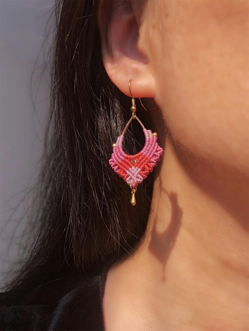 May include: A handmade macrame earring with a gold-colored hook and a teardrop-shaped design. The earring features a gradient of pink and red threads, with a small gold bead and a dangling gold teardrop charm.