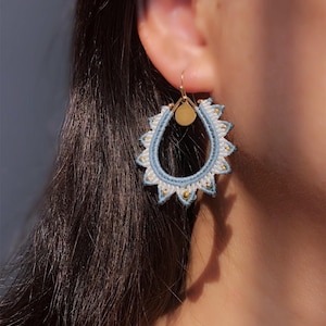 Può includere: Un orecchino a forma di goccia con ricamo in filo azzurro e bianco a raggiera. Il design include piccole perline color oro lungo il bordo interno e un disco in metallo dorato nella parte superiore, sospeso a un gancio color oro.