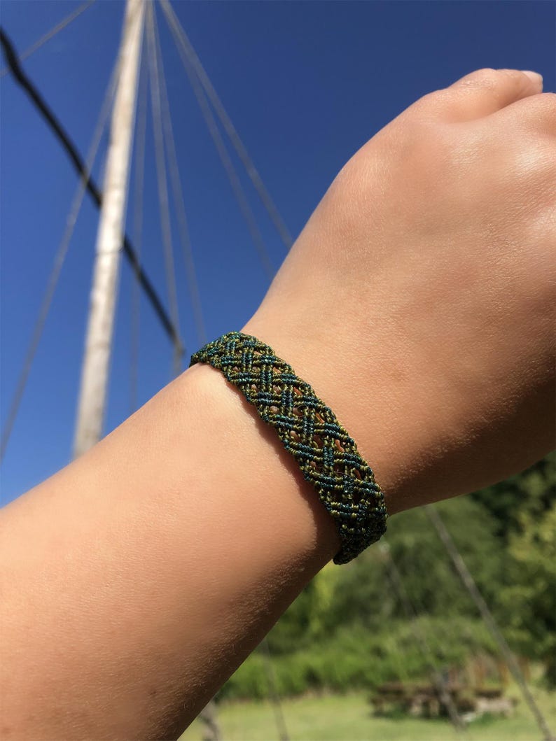 K&ouml;nnte beinhalten: Ein geflochtenes Armband mit einem komplexen Gittermuster aus dunkelblaugr&uuml;nen und olivgr&uuml;nen Kordeln. Das Accessoire besteht aus mehreren miteinander verflochtenen F&auml;den, die ein sich wiederholendes, rautenf&ouml;rmiges geometrisches Design bilden.