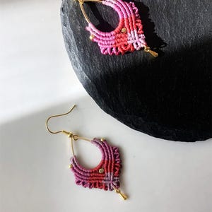 May include: A pair of handmade macrame earrings in shades of pink and red. The earrings feature a gold-colored hook and small gold accents. The macrame design is intricate and detailed, with a unique shape.
