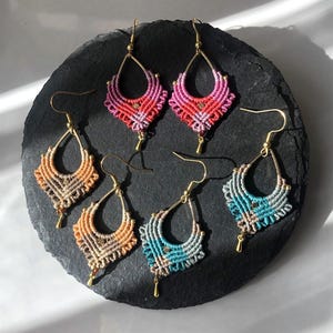 May include: Six pairs of handmade macrame earrings in various colors, including pink, orange, and teal. Each earring features a teardrop-shaped design with gold-colored hooks and accents. The earrings are displayed on a dark, textured surface.
