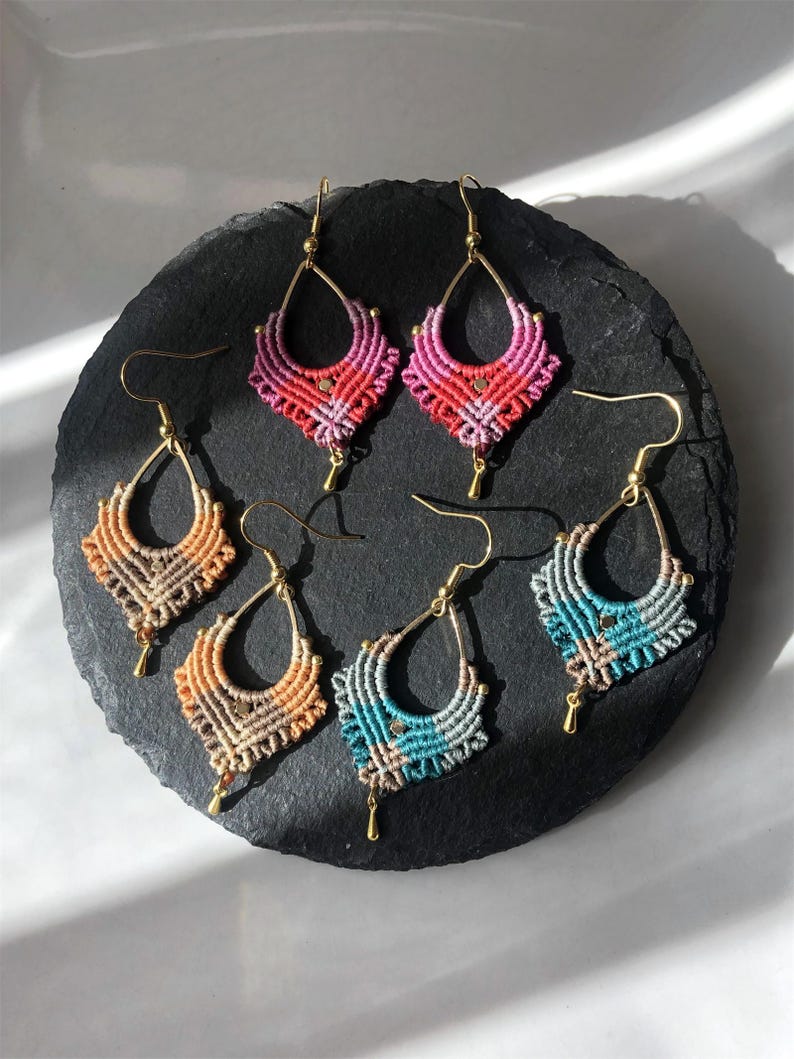 May include: Six pairs of handmade macrame earrings in various colors, including pink, orange, and teal. Each earring features a teardrop-shaped design with gold-colored hooks and accents. The earrings are displayed on a dark, textured surface.