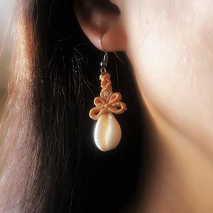 May include: A single earring featuring a natural seashell pendant. The earring has a tan macrame design with a small, white shell. The earring is suspended from a silver-colored hook.