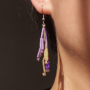 Puede incluir: Primer plano de un pendiente de macramé con un gancho plateado. El pendiente presenta hebras de cordón lavanda, dorado y morado, acentuadas con pequeñas cuentas. El diseño es intrincado y hecho a mano, mostrando un estilo bohemio.