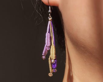 May include: Close-up of a macrame earring with a silver hook. The earring features strands of lavender, gold, and purple cord, accented with small beads. The design is intricate and handmade, showcasing a bohemian style.
