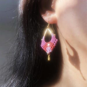 May include: A handmade macrame earring in shades of pink and red, with a gold-colored hook and a small gold bead at the bottom. The earring is an intricate design, with a geometric shape.