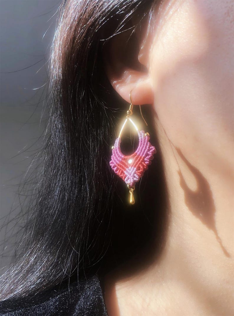 May include: A handmade macrame earring in shades of pink and red, with a gold-colored hook and a small gold bead at the bottom. The earring is an intricate design, with a geometric shape.
