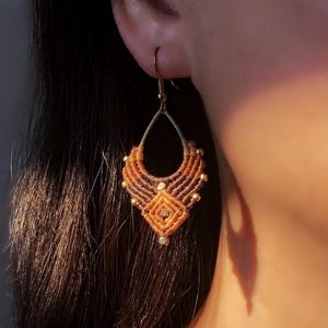 Könnte beinhalten: Handgefertigte Makramee-Ohrringe in Braun- und Goldtönen. Die Ohrringe haben eine Tropfenform mit aufwendigen Knoten und kleinen goldenen Perlen. Das Design beinhaltet ein zentrales Rautenmuster. Sie hängen an goldfarbenen Ohrhaken.