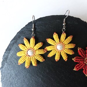 May include: A pair of yellow flower earrings with a white pearl center. The earrings are made with a macrame technique and have silver hooks.