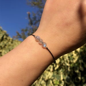 May include: A black cord bracelet featuring three round, light-colored beads. The bracelet is worn on a wrist, with a background of green foliage and a blue sky.